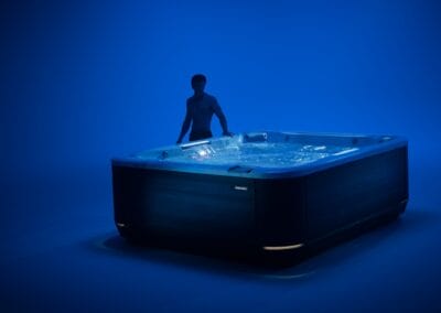 Man standing beside a large, dark wood hot tub in a blue-lit studio setting