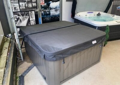 Large outdoor hot tub with a gray protective cover on display in a showroom, next to a smaller spa with jets visible in the background
