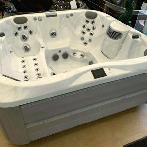 Large white hot tub with numerous jets and built-in seats, shown in an indoor showroom display with a gray exterior paneling side.