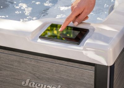Person taps a touchscreen control on the edge of a white hot tub, water visible in the background.