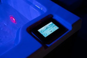 Touchscreen control panel mounted on the edge of a blue-lit hot tub, displaying settings on its screen