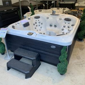 Outdoor hot tub with white interior and black cabinet, two-step black stairs in front, showroom display with signage and greenery nearby,