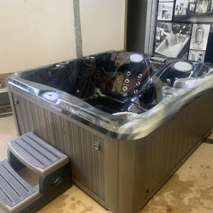 Large black outdoor hot tub with multiple jets and built-in seating, placed in a showroom with gray steps nearby.
