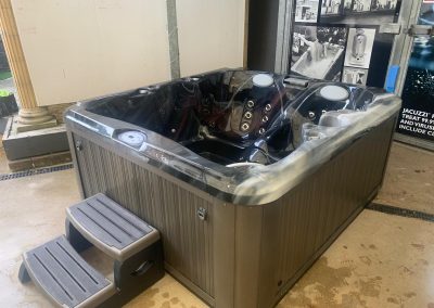 Large black outdoor hot tub with multiple jets and built-in seating, placed in a showroom with gray steps nearby.