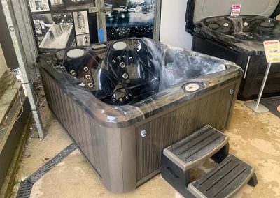 Indoor showroom hot tub with dark panels and plastic cover, built-in jets visible, small black steps nearby.