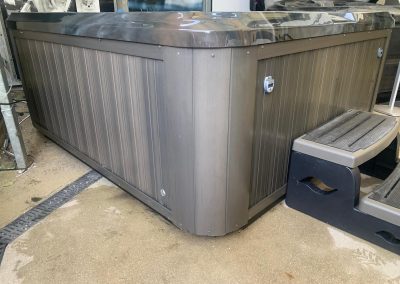 Outdoor hot tub with dark brown paneled sides and stainless steel top, placed on a concrete surface with a small step stool nearby.