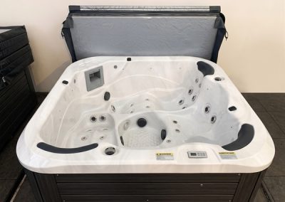 White rectangular hot tub with multiple jets and built-in headrests, open lid in a showroom setting with a dark wood exterior paneling nearby.