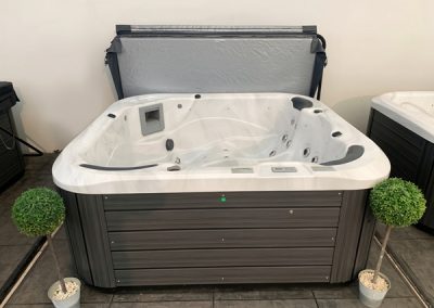 White acrylic hot tub with a dark gray cabinet and an open lid in a showroom, flanked by two small potted topiary trees. jets visible inside the tub.