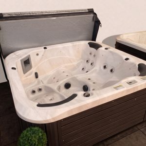 White acrylic hot tub with multiple jets and built-in seating, on a wooden deck with a lid open and potted plants nearby.