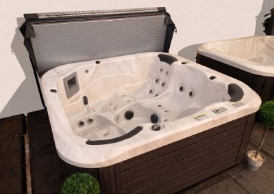 White acrylic hot tub with multiple jets and built-in seating, on a wooden deck with a lid open and potted plants nearby.